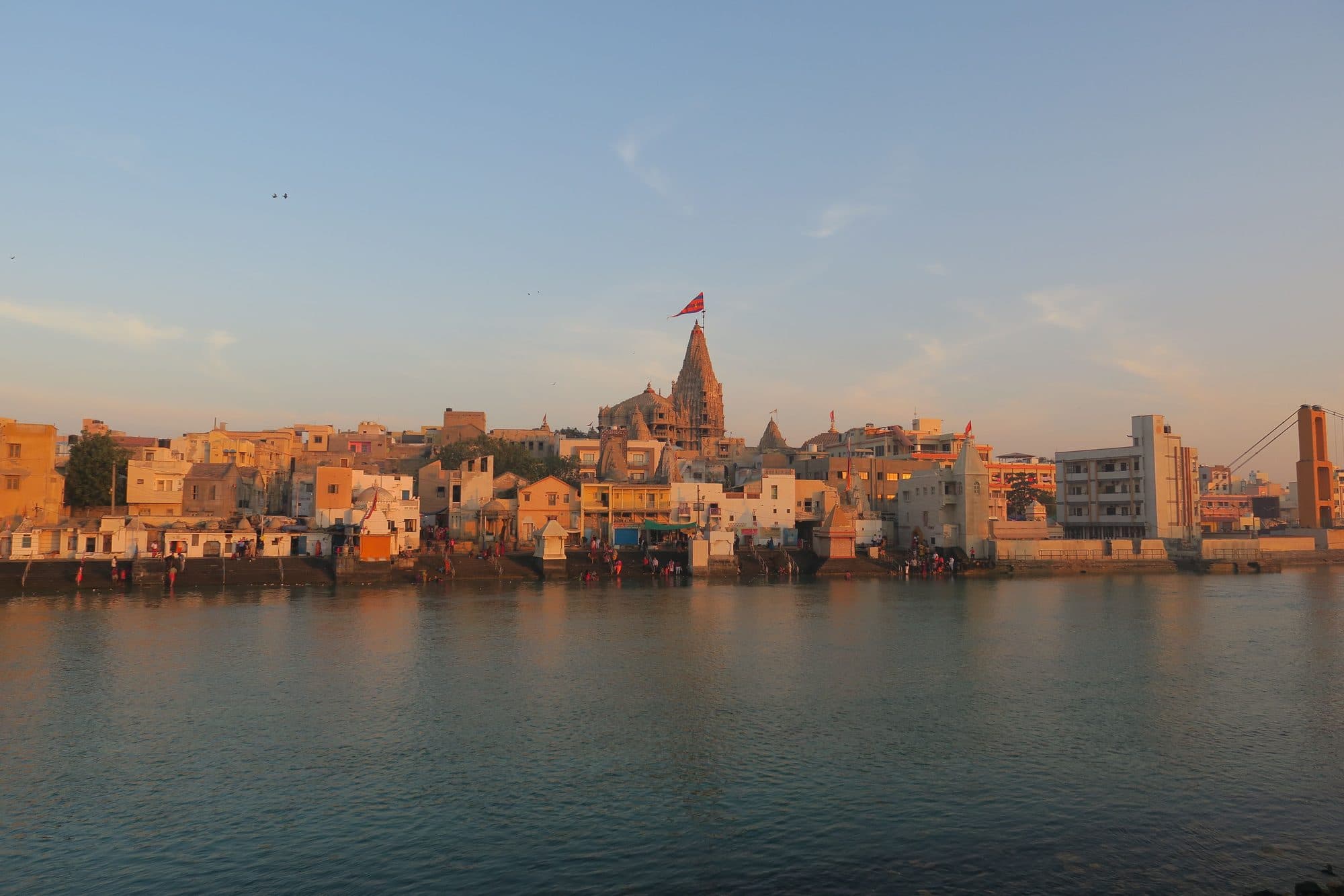 The spire and flag of Dwarkadhish Temple rising above the old town of Dwarka, seen across the Gomti river and its bathing ghats in warm evening light