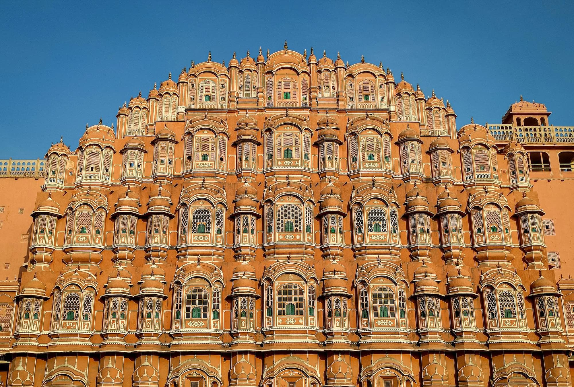 Front view of Hawa Mahal's pink sandstone facade in Jaipur, its tiers of latticed jharokha windows lit by warm sun under a clear blue sky