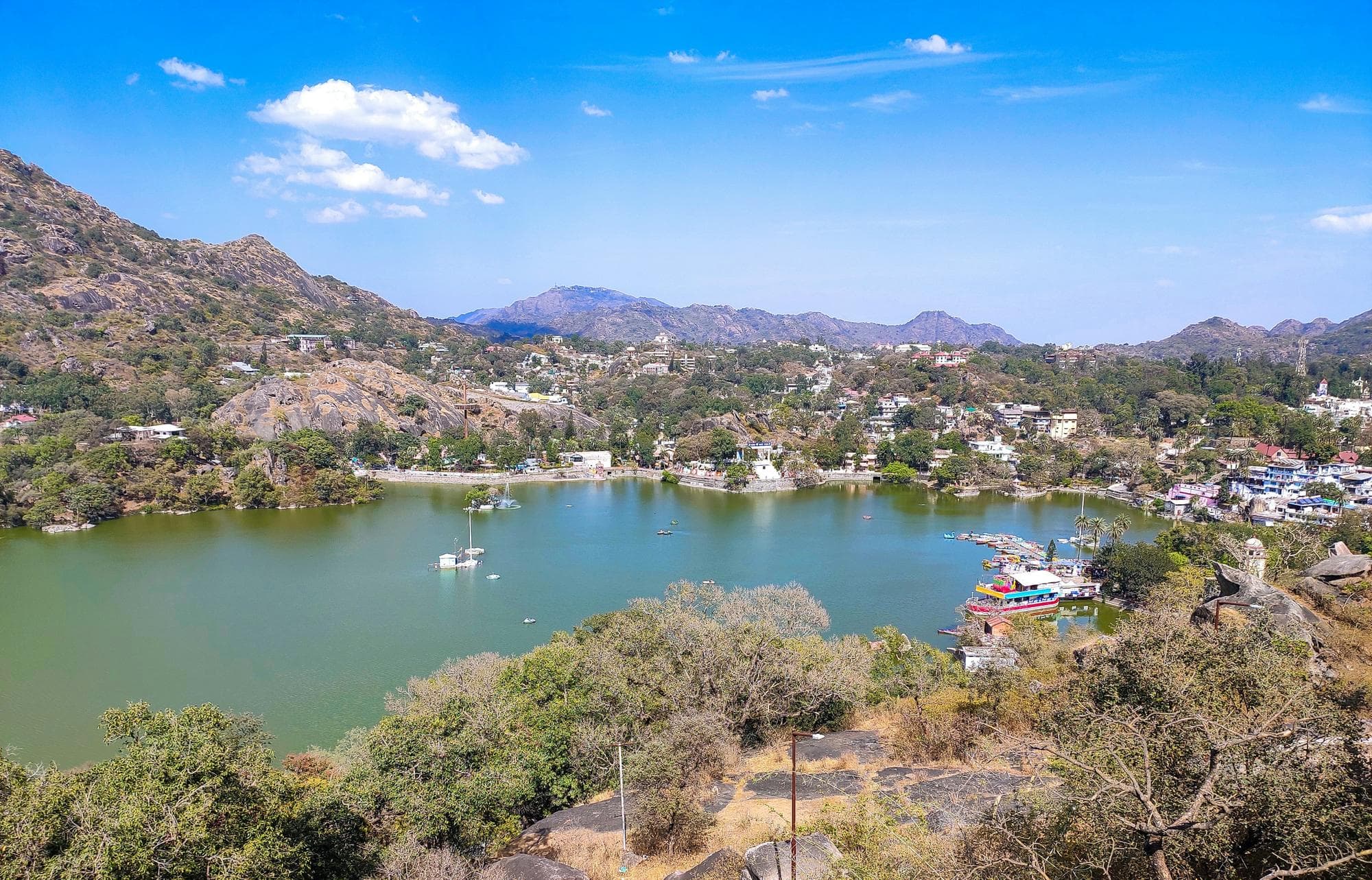 Elevated view over Nakki Lake in Mount Abu on a sunny afternoon, with boats on the green water, the hill town along its shore and the Aravalli hills behind