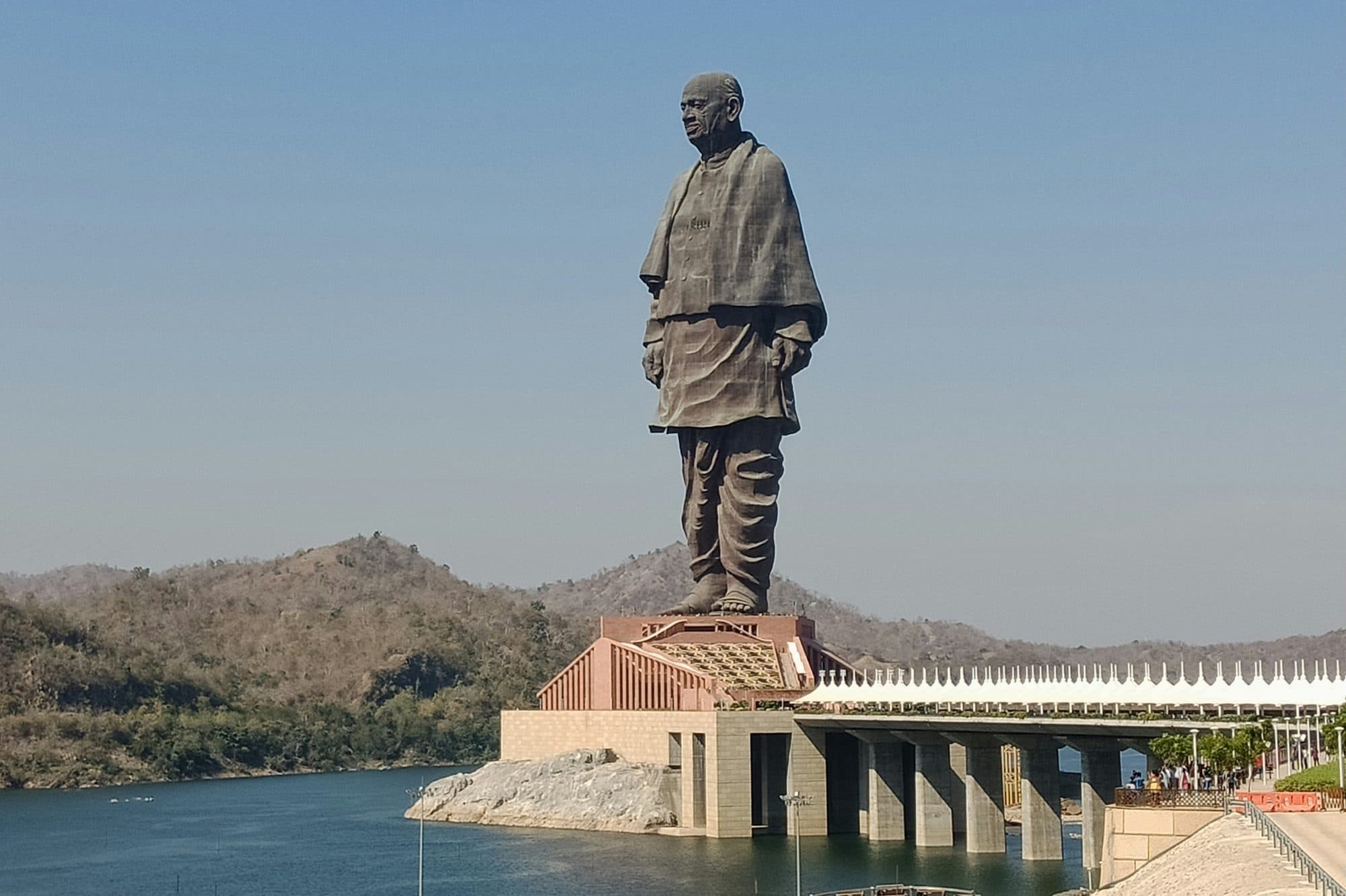 The Statue of Unity on its island in the Narmada river near Kevadia, Gujarat, with dry hills behind and the covered access bridge on the right