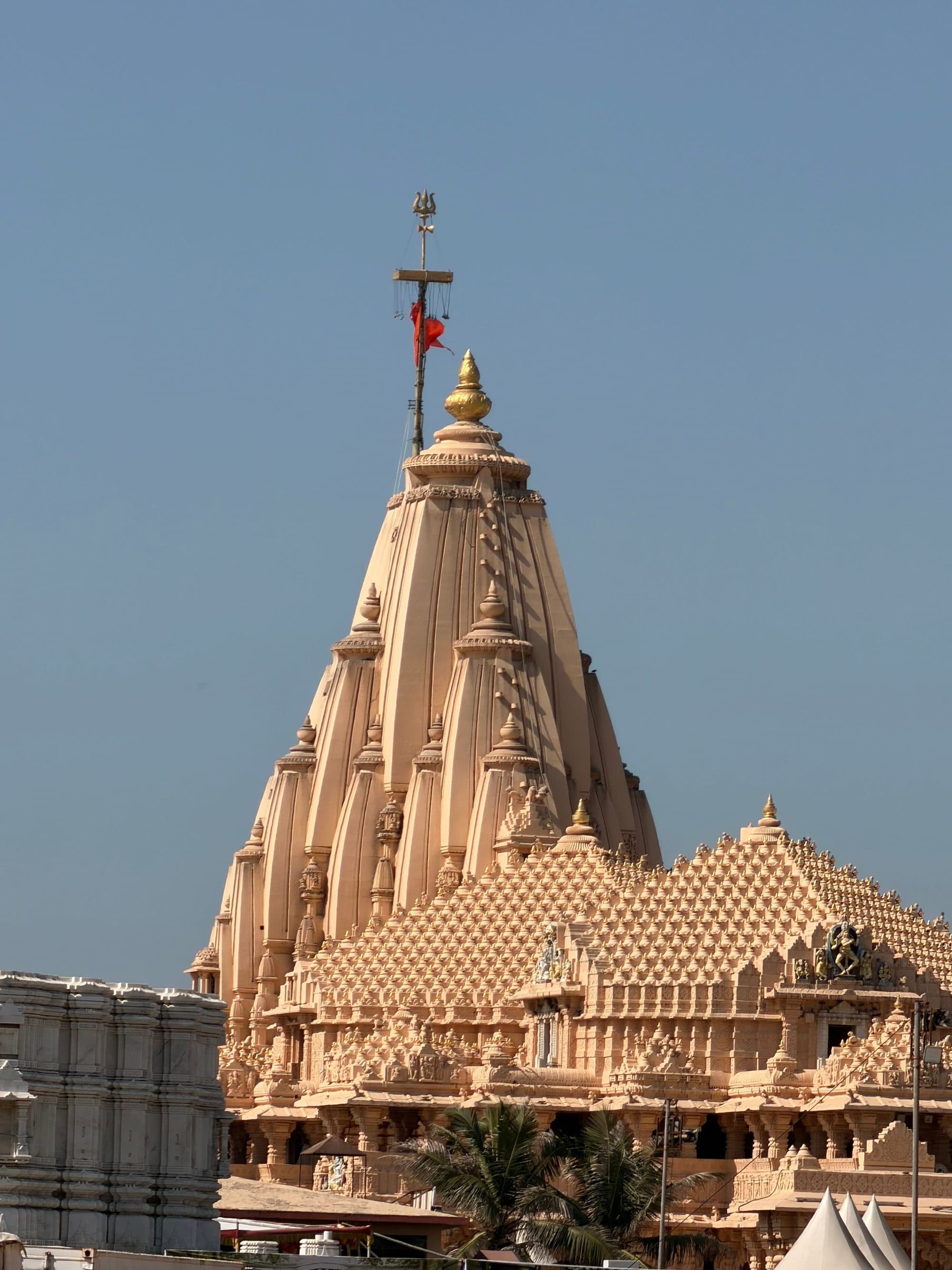 The carved sandstone spire of Somnath Temple in Gujarat, topped by a golden finial and a flag, rising above the temple's pyramidal hall roof against a clear blue sky